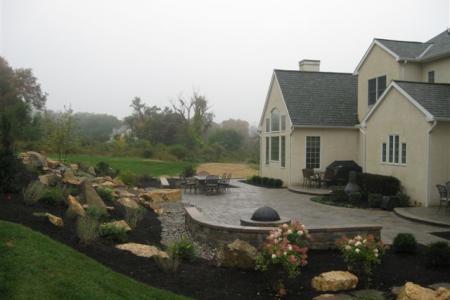 A gorgeous backyard in Malvern, Pa includes a paver patio, pondless waterfall, plantings, fire pit, witting wall and lighting.